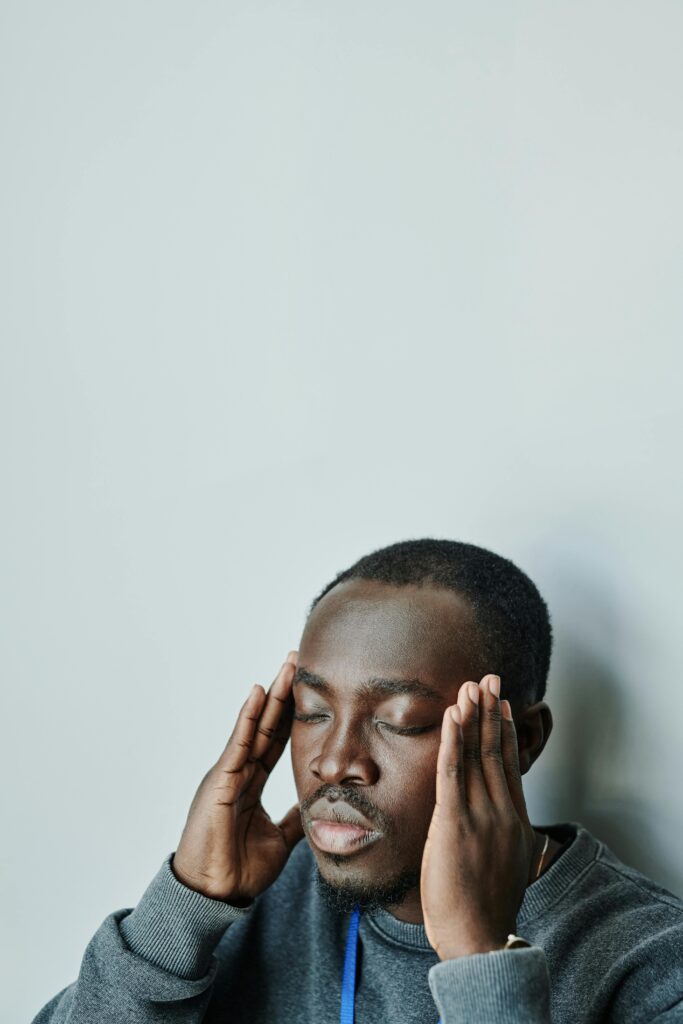 A black man with closed eyes concentrates and meditates indoors wearing gray long sleeves.