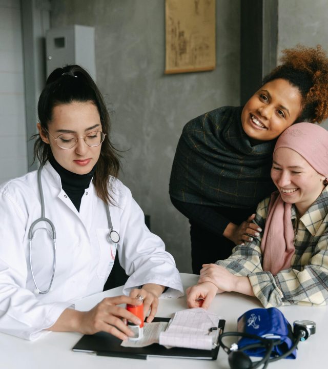 A doctor consults a cancer patient and her supportive friend in a warmly lit clinic room.